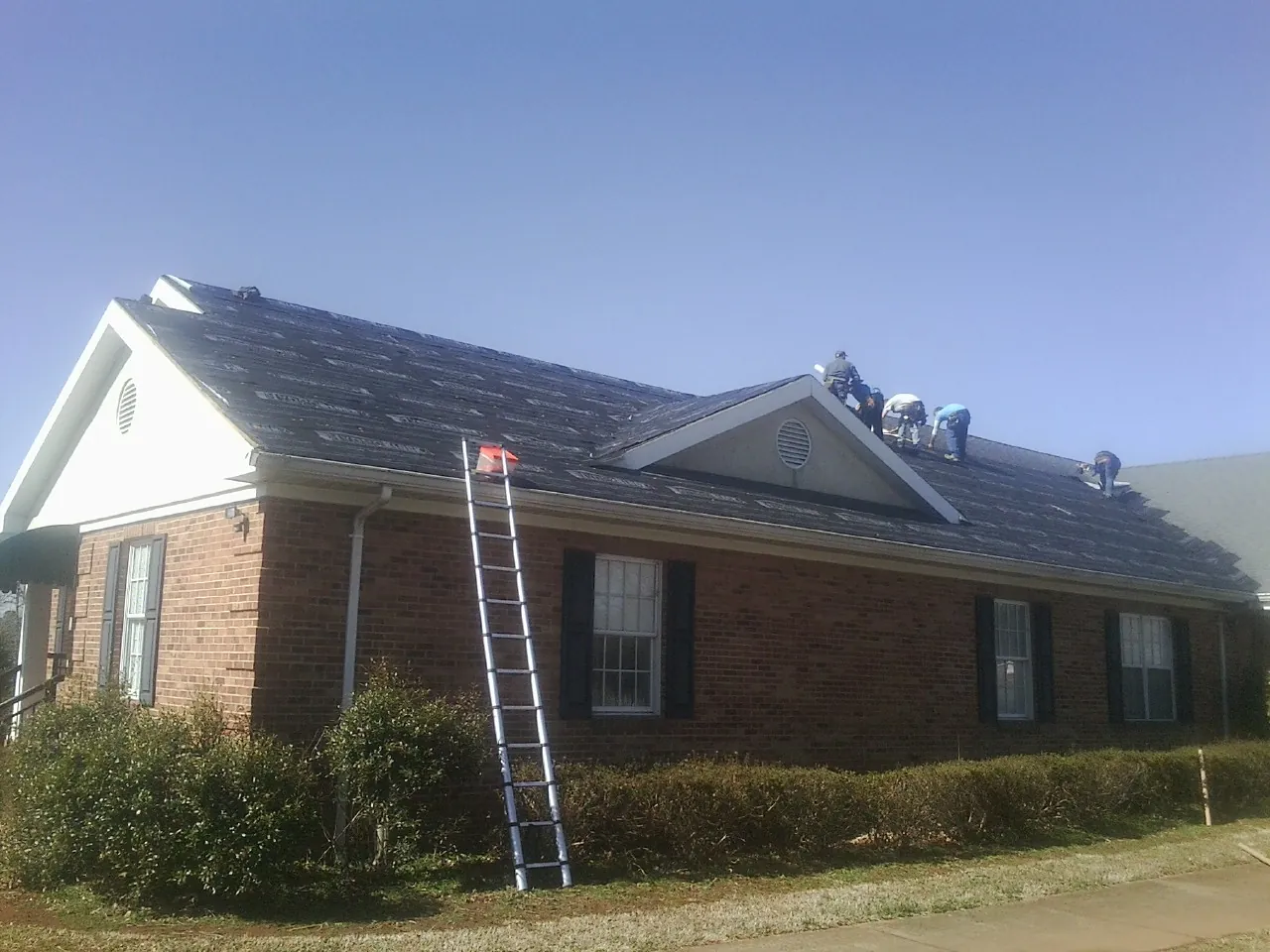 Crew tearing shingles off a brick ranch roof with a ladder set — Metal Roof Repair work by our crew in Gilson