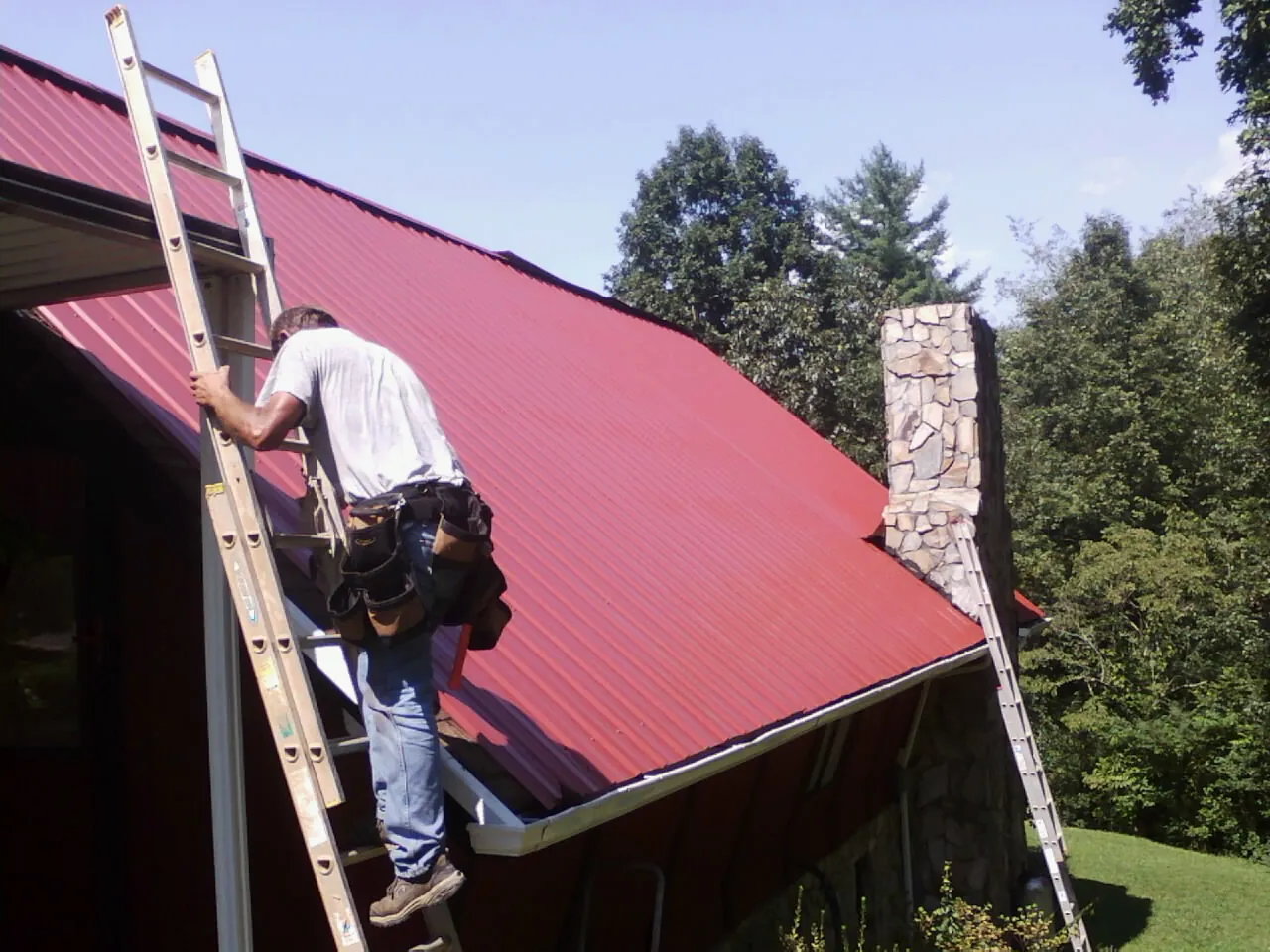 Roofer on a ladder beside a red metal roof and stone chimney — Soffit & Fascia Repair by Roofing Brothers in Gilson